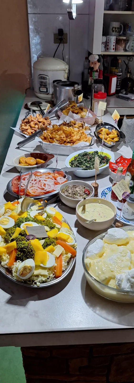 Mesa comprida posta com dezenas de travessas: salada de tomate com cebola, couve, saladas de folhas com ovo e manga, e um caldo cremoso